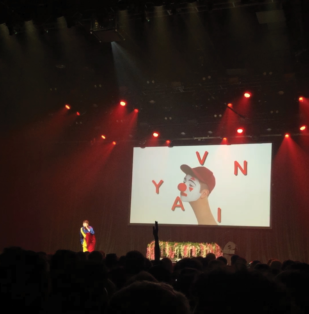 This is a photo of Yavin singing onstage while wearing a clown costume in front of a screen with his name on it.